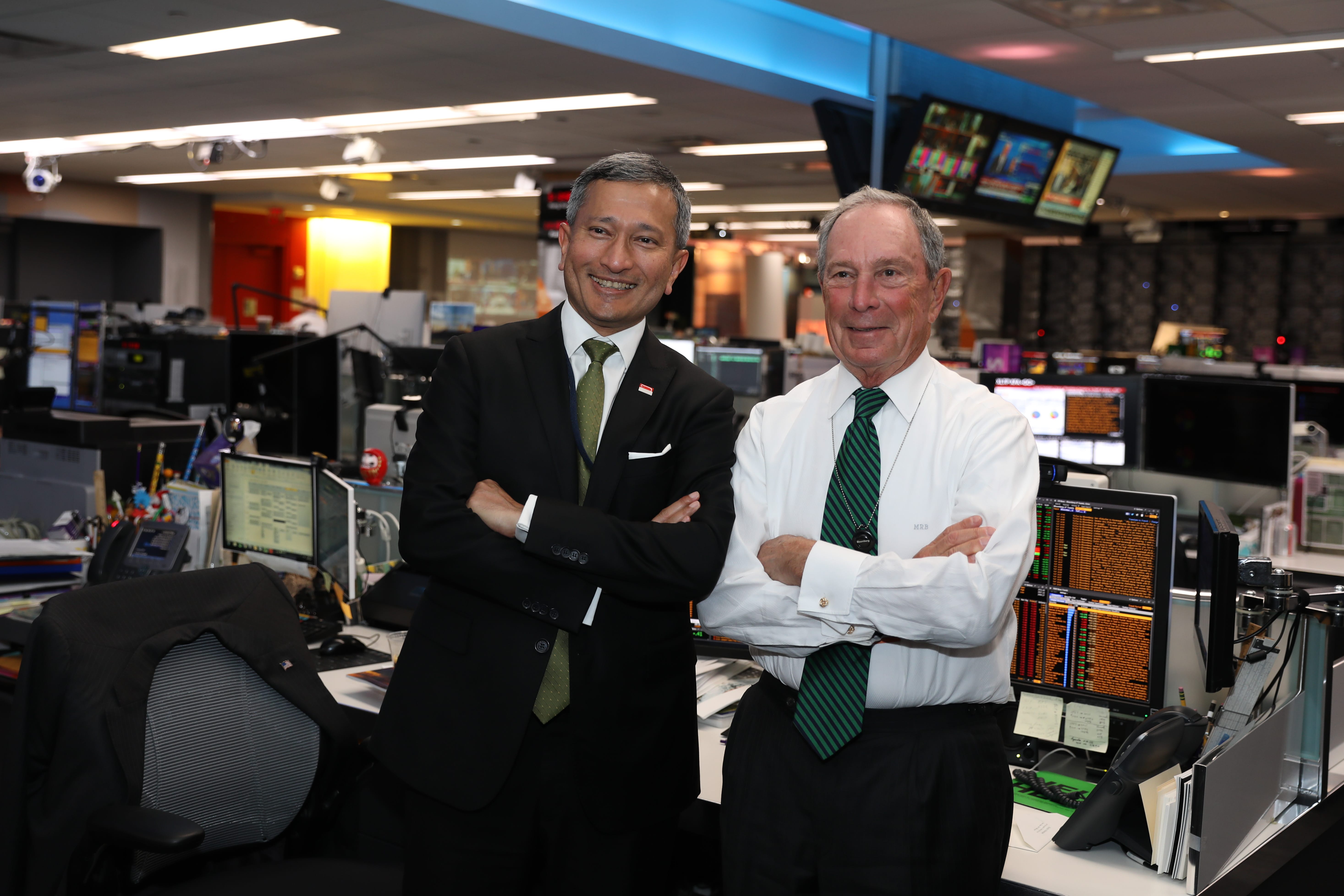 Two men stand with arms crossed in a busy Bloomberg newsroom. Michael Bloomberg is on the right.